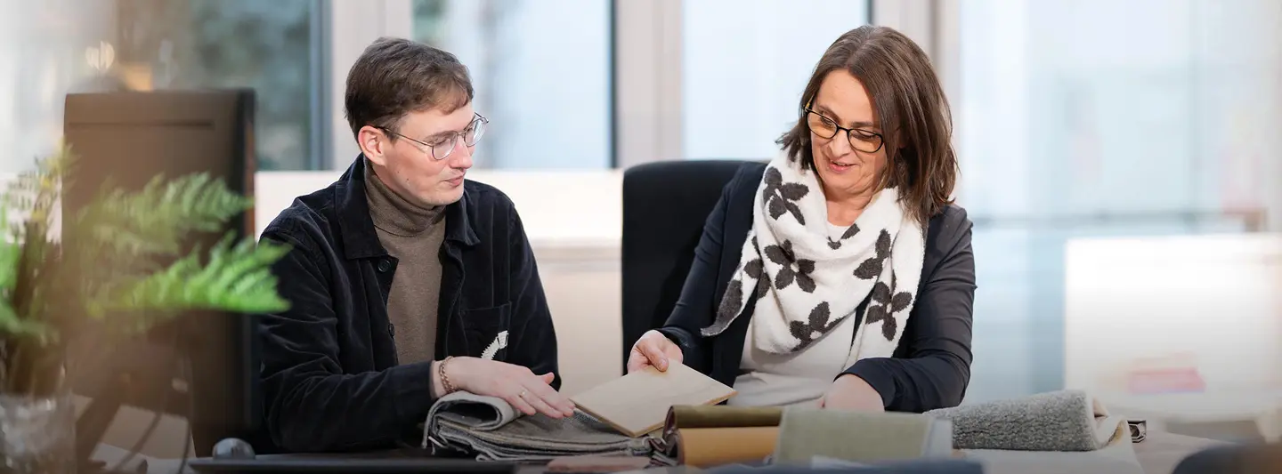 Zwei Personen sitzen am Tisch und betrachten Stoffmuster und Holzprobe, Ordner und Materialien liegen vor ihnen im Büro